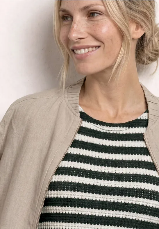Femme blonde souriante de profil, portant un top à rayures noir et blanc et un blazer beige.