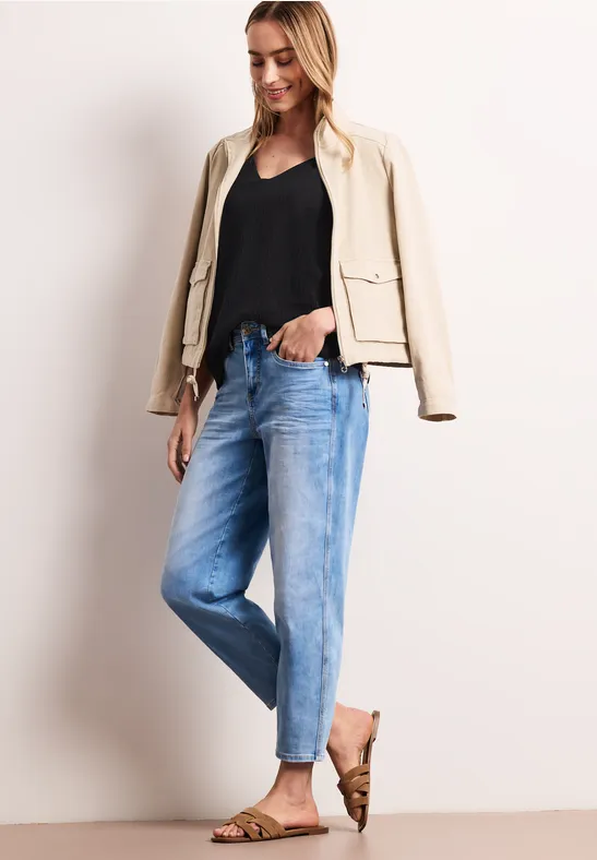 Vrouw met zwarte top, beige jasje, lichtblauwe jeans en bruine sandalen voor neutrale achtergrond.