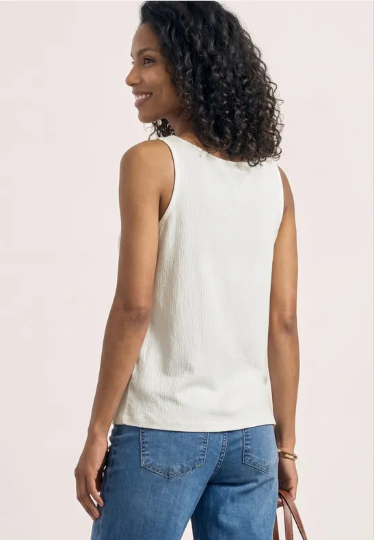 Person von hinten mit lockigem Haar, weißem ärmellosen Top und blauer Jeans vor neutralem Hintergrund.