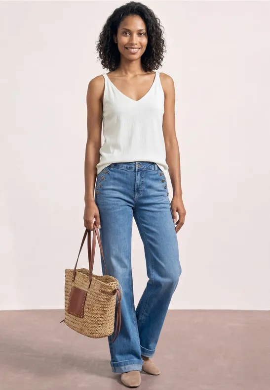 Eine Frau trägt ein weißes Top, weite blaue Jeans und hält eine geflochtene Korbtasche.