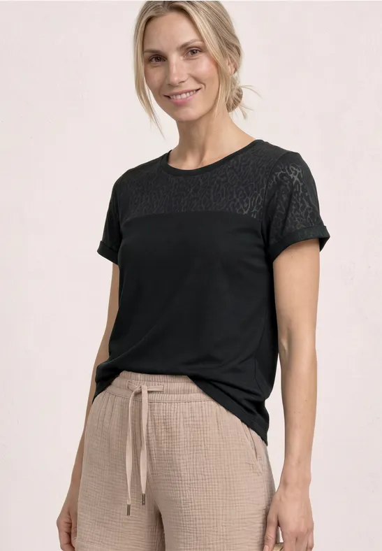 Frau vor hellem Hintergrund trägt schwarzes T-Shirt mit Spitzenmuster und beige Hose mit Kordelzug