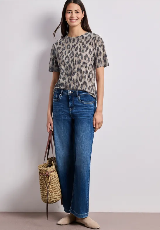Frau trägt kurzärmeliges Leopard-Shirt, weite Jeans, Basttasche und flache Schuhe.