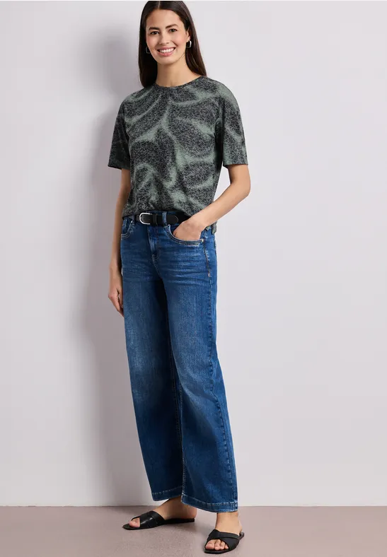 Frau trägt bedrucktes grünes T-Shirt, weite blaue Jeans, schwarzen Gürtel und Sandalen.