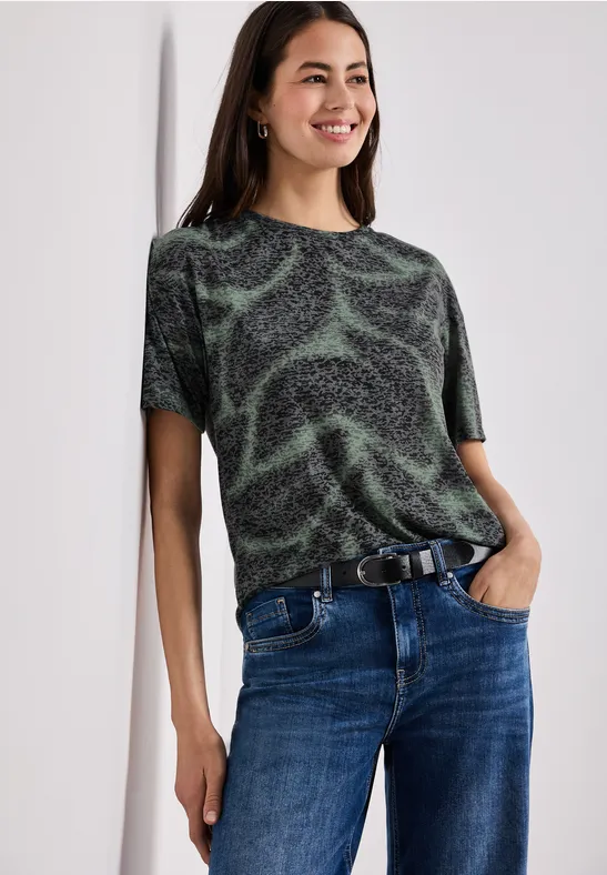 Frau in schwarzem T-Shirt mit grünen Wirbelmustern und Jeans lehnt lässig an der Wand.