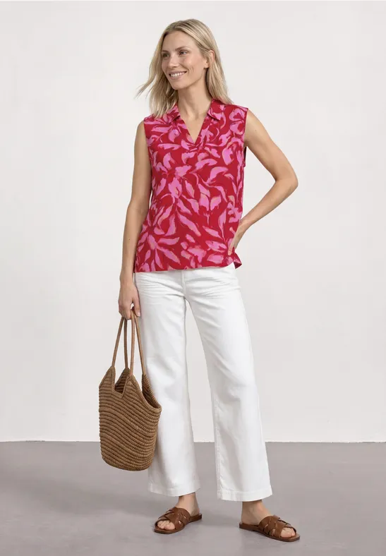 Une femme porte un haut rose à motif, un pantalon blanc, des sandales marron et un sac en paille tressée.