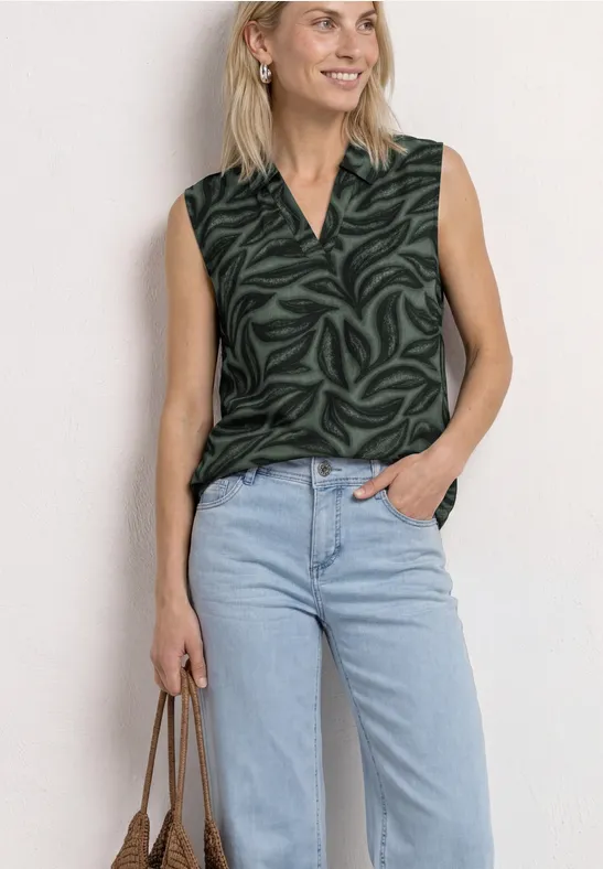 Vrouw in mouwloze donkergroene blouse met patroon en V-hals, lichte jeans en een handtas.