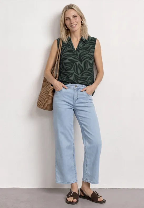 Vrouw met mouwloos groen bladprinttop, lichtblauwe hoge taille jeans, bruine sandalen en rieten tas.