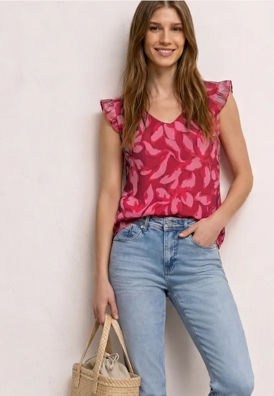 Frau in pinker gemusterter Bluse mit Rüschenärmeln, hellblauen Jeans und kleiner Stroh-Tasche vor hellem Hintergrund.