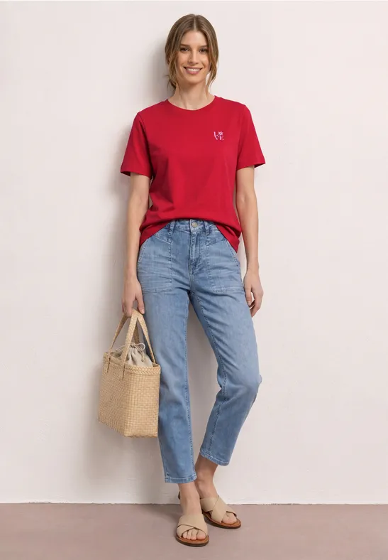 Vrouw poseert tegen neutrale achtergrond in rood T-shirt, losse spijkerbroek, platte sandalen en rieten tas