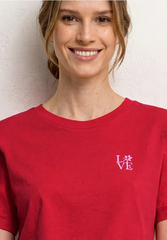 Persoon in een rood T-shirt met klein wit 'LOVE'-logo op de borst, voor een witte muur.