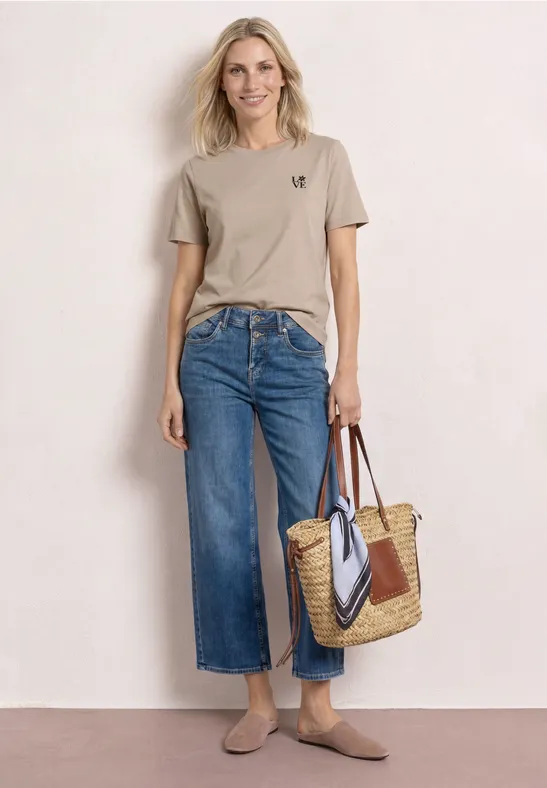 Frau vor hellem Hintergrund mit beigem T-Shirt, weitem Jeans-Culotte, Basttasche und flachen Schuhen.