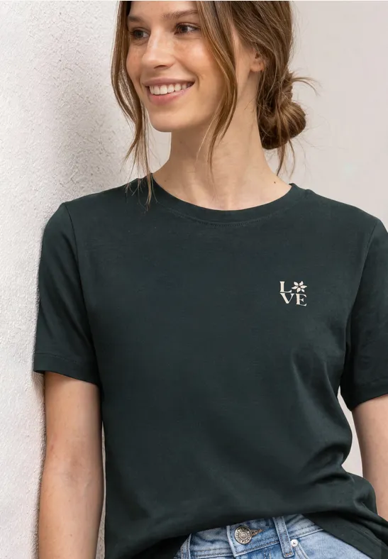 Junge Frau trägt schwarzes T-Shirt mit kleinem weißen „love“-Schriftzug, vor hellem Hintergrund.