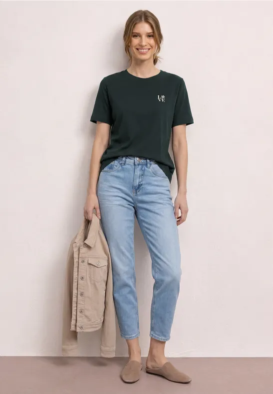 Frau in schwarzem t-Shirt, hellen Jeans, beigen Schuhen, hält beige Jacke, vor neutralem Hintergrund.