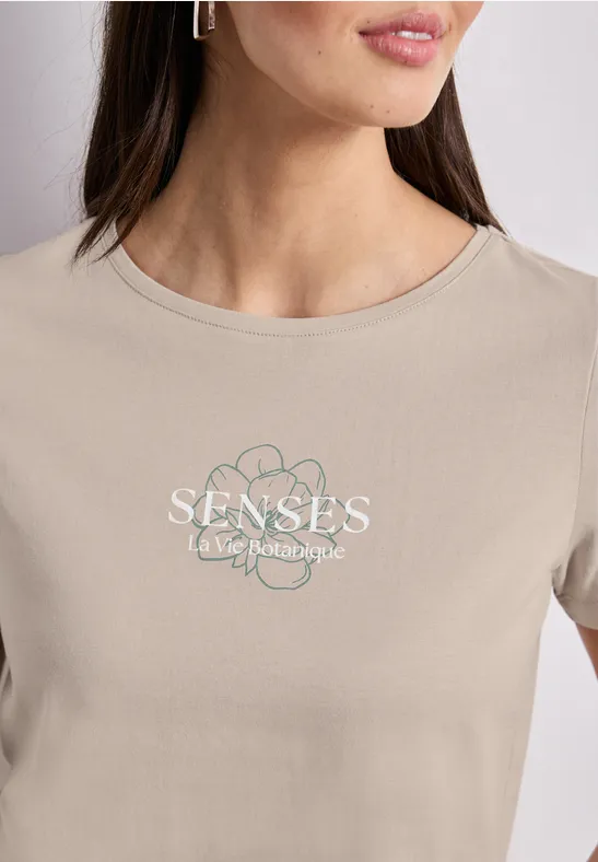 Close-up van een vrouw in een beige t-shirt met bloemmotief en tekst 'SENSES - take back your style'
