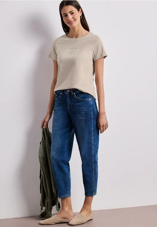 Vrouw draagt beige T-shirt en blauwe jeans, houdt een groene jas vast, casual outfit.