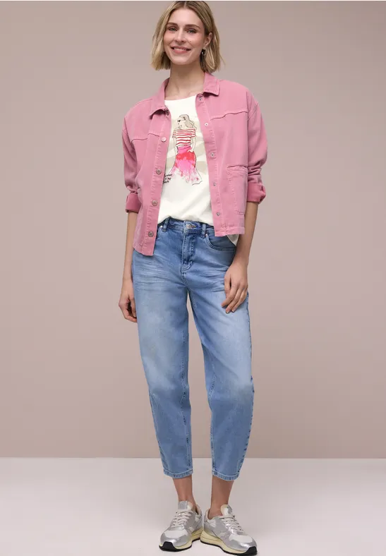 Model draagt roze spijkerjack, wit T-shirt met print, losse blauwe jeans en sneakers tegen neutrale achtergrond.