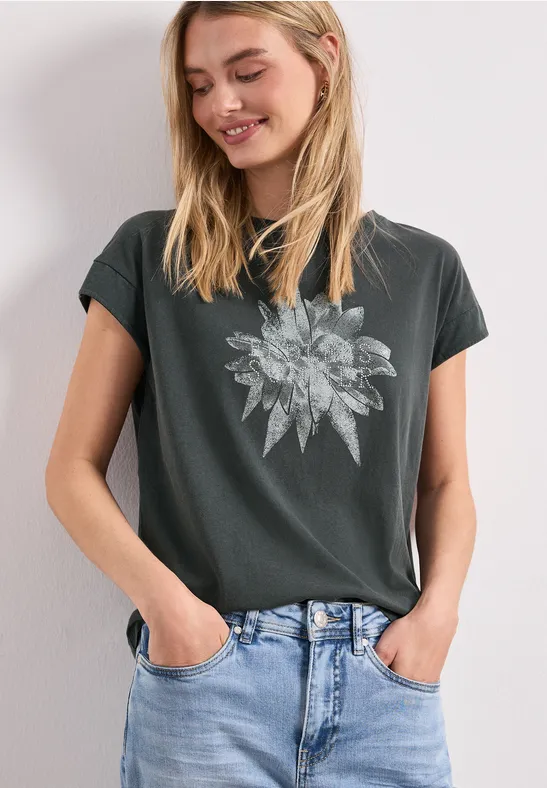 Frau in dunklem T-Shirt mit floralem Blattmotiv und blauen Jeans, Hände in den Hosentaschen.