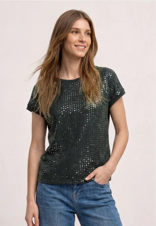 Une femme porte un t-shirt vert à sequins et un jean bleu, posant devant un fond clair.