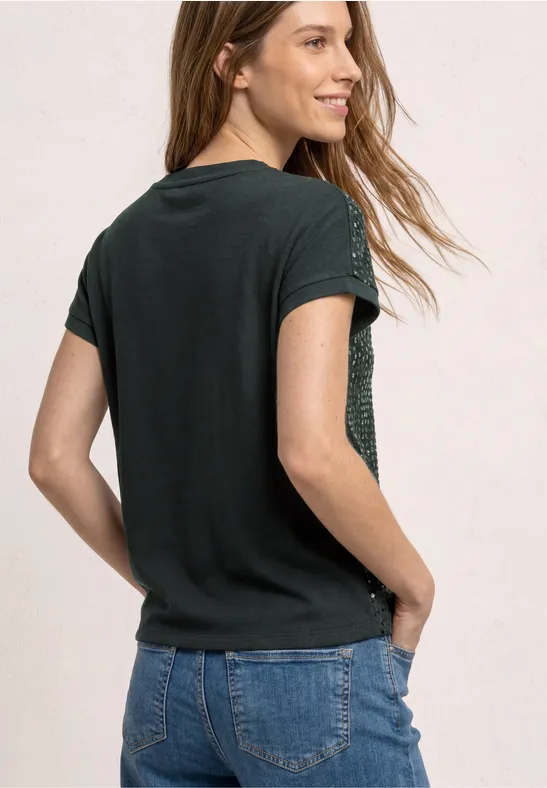 Jeune femme de dos, en t-shirt noir et jean, mains dans les poches, devant un fond clair.