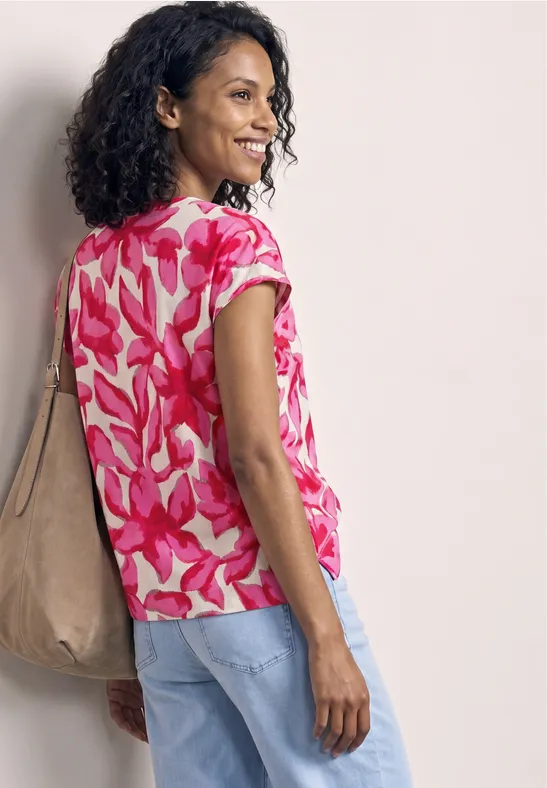 Frau in floraler rosa Bluse, hellen Jeans und beiger Tasche steht seitlich vor neutralem Hintergrund.