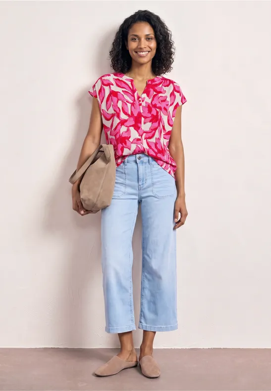 Frau in pink gemusterter Bluse, hellblauen Culotte-Jeans, beigem Shopper und Loafers.