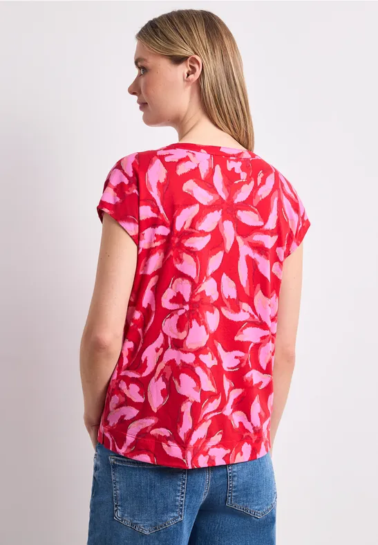 Frau von hinten in roter Bluse mit rosa Blattmuster und blauer Jeans.