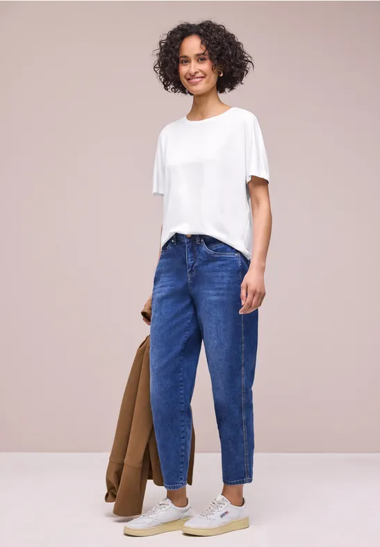 Iemand met wit T-shirt en blauwe spijkerbroek, houdt een bruine jas vast voor lichte achtergrond.