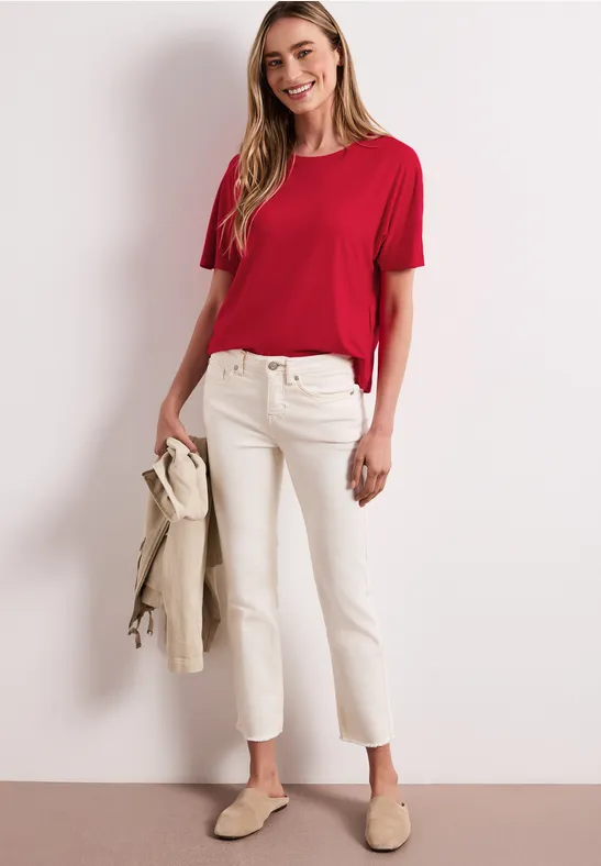 Vrouw in rode T-shirt, witte broek en beige instappers, met een beige jasje in de hand.