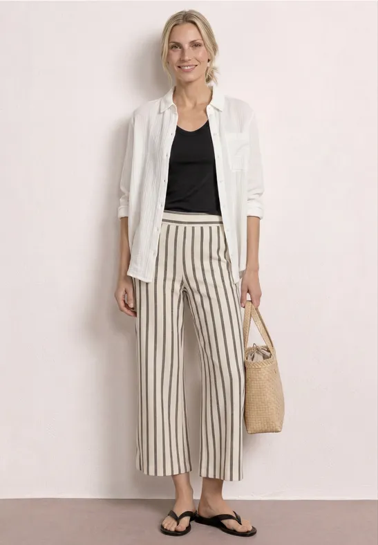 Vrouw met wit hemd over zwart topje, gestreepte wijdvallende broek, slippers en gevlochten tas op neutrale achtergrond.