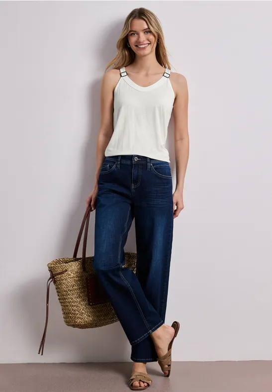 Vrouw in wit topje, donkere jeans, espadrilles en strooien tas voor neutrale achtergrond.