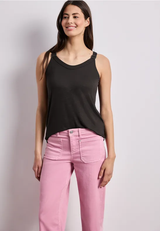 Een vrouw in een zwarte tanktop en roze spijkerbroek staat voor een neutrale achtergrond.