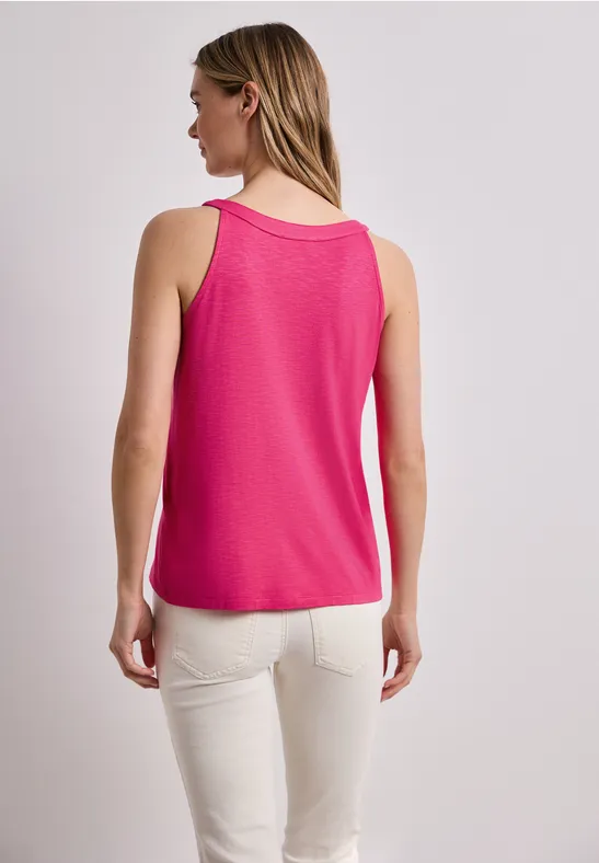 Vrouw van achteren in een felroze mouwloze top en lichte broek tegen neutrale achtergrond.