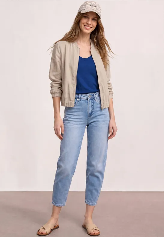 Frau in beiger Jacke, dunklem Top und hellblauen Jeans, flache Sandalen