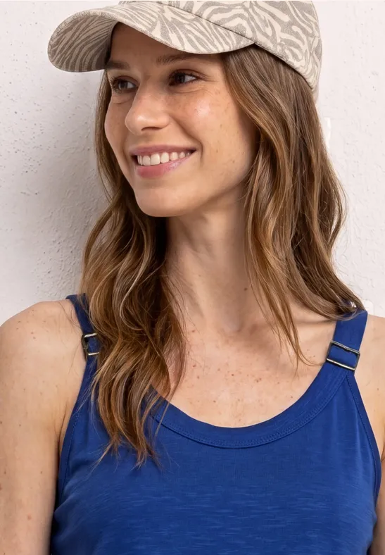 Eine Person trägt ein blaues Trägershirt und eine gemusterte Kappe vor grauem Hintergrund.
