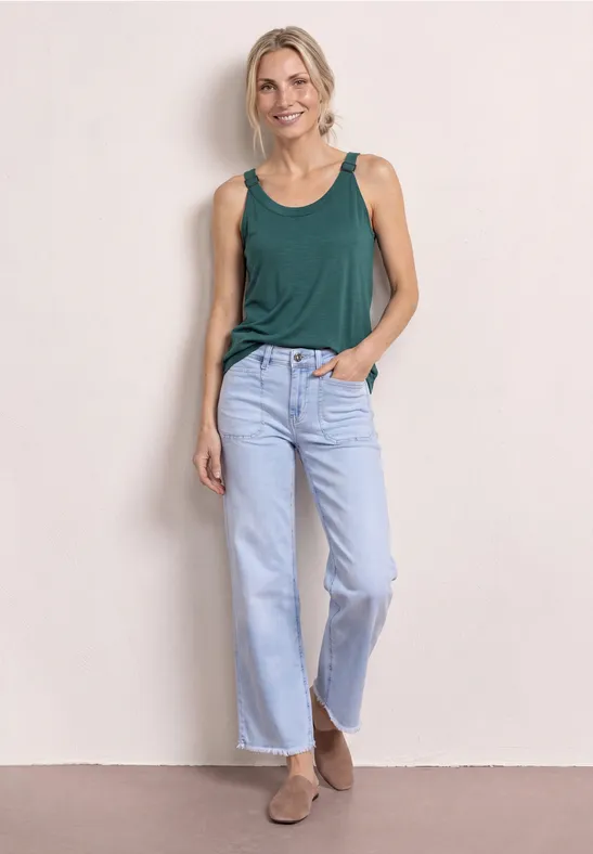 Frau lehnt in grünem Top, blauen Jeans und braunen Stiefeln lässig an einer Wand.