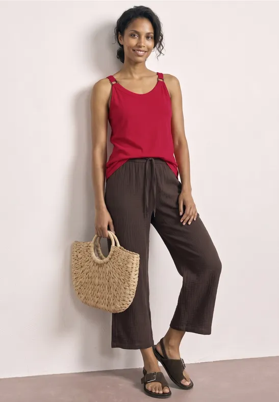 Vrouw met rood mouwloos topje, bruine wijde broek, zwarte sandalen en gevlochten strooien tas.