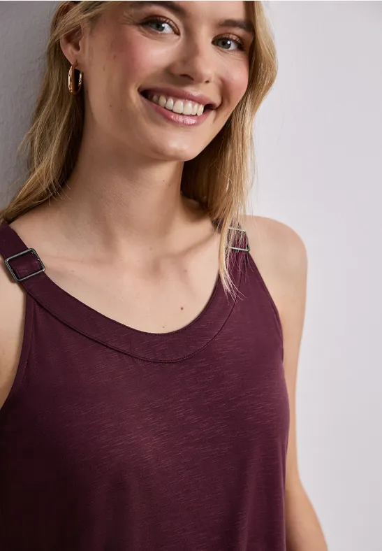 Vrouw in bordeauxroze mouwloze top met metalen schouderdetail en gouden oorbellen voor een neutrale achtergrond.