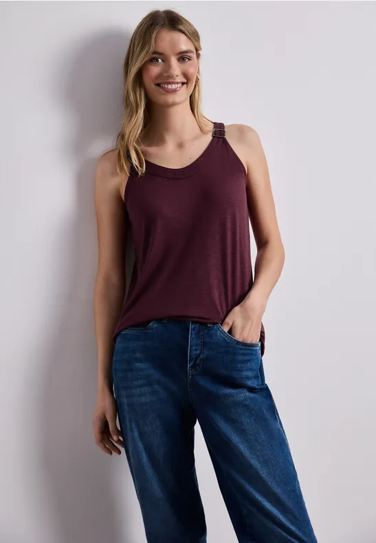 Vrouw staat ontspannen voor een witte muur, draagt een bordeaux top en een blauwe spijkerbroek.