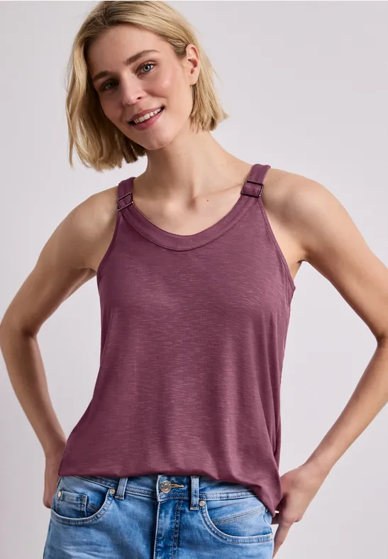 Eine Frau mit hellem Haar trägt ein ärmelloses weinrotes Top und blaue Jeans vor hellem Hintergrund.