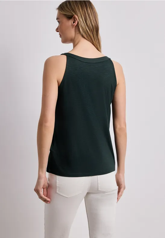 Vrouw van achteren in een groen hemdje en witte broek tegen een neutrale achtergrond.