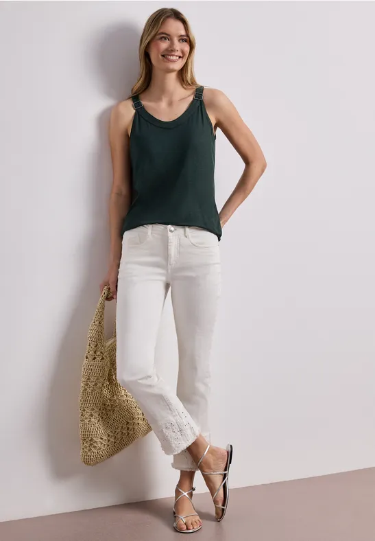 Vrouw in groen hemdje, witte jeans, sandalen en rieten tas leunend tegen een witte muur.