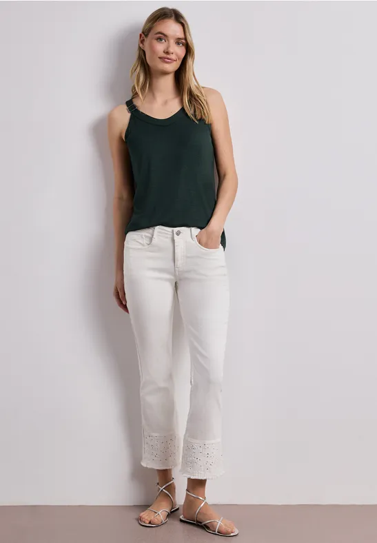 Jonge vrouw in een zwart topje, witte broek met kant en sandalen tegen lichte achtergrond.