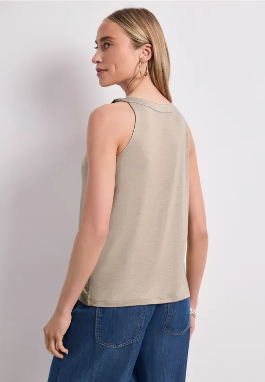 Rückenansicht einer Frau in beigem ärmellosem Top und blauen Jeans vor hellem Hintergrund.