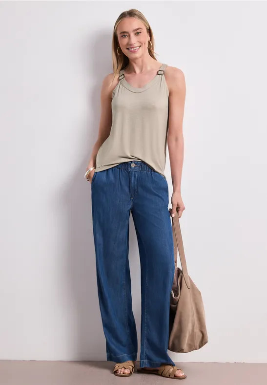 Frau steht in lockerer Pose, trägt beige Top, weite blaue Jeans und eine große beigefarbene Tasche vor weißem Hintergrund.
