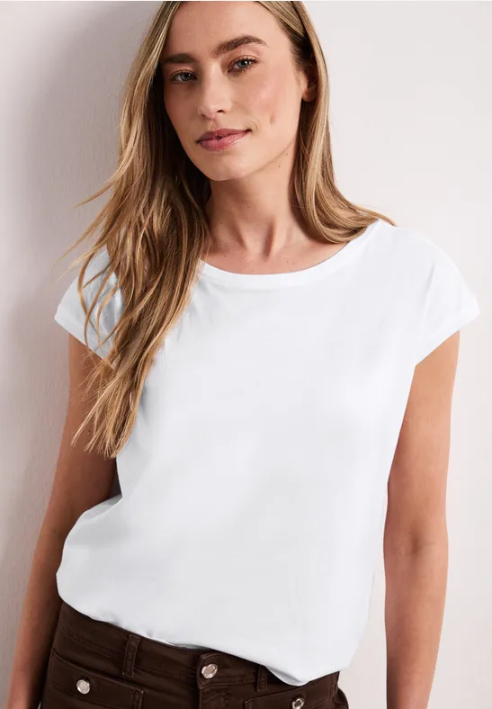 Vrouw in wit T-shirt en bruine broek voor een lichte achtergrond.