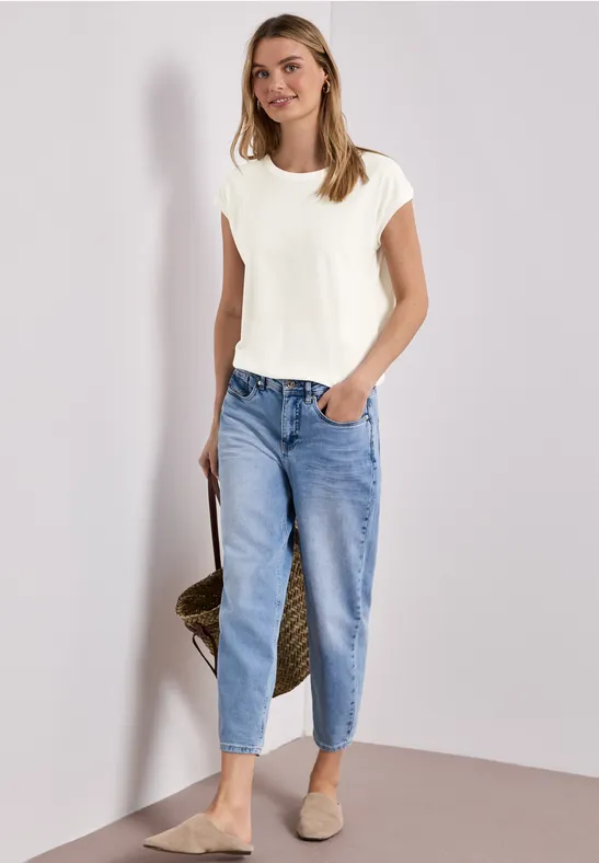 Vrouw in wit T-shirt en losse blauwe jeans met strozak, ontspannen pose tegen lichte achtergrond.