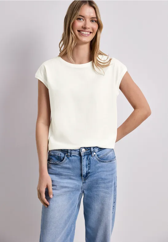 Een vrouw draagt een wit T-shirt met korte mouwen en blauwe jeans, poserend voor een neutrale achtergrond.