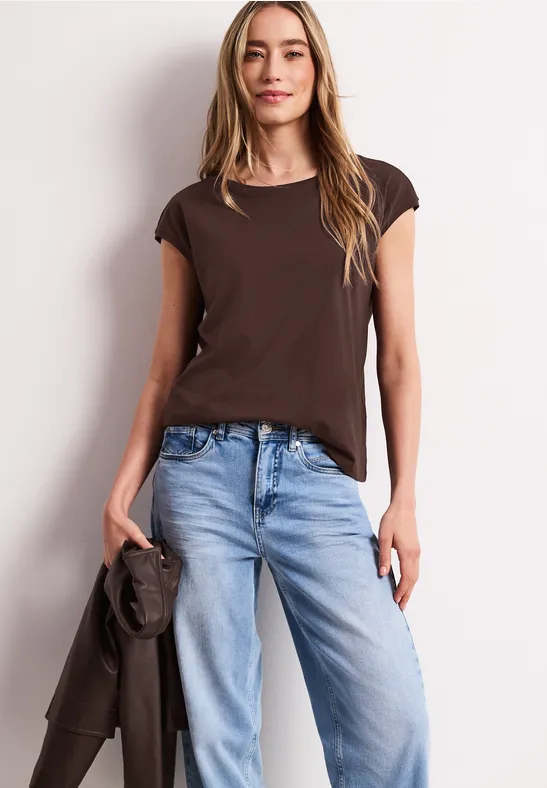 Femme en t-shirt marron et jean bleu clair, tenant une veste, posée devant un fond blanc.