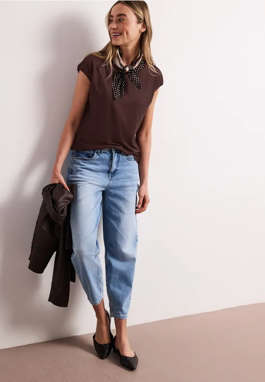 Femme en haut marron, foulard, jean clair raccourci, ballerines noires, tenant un manteau.