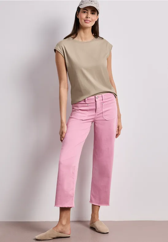 Frau trägt beige ärmellose Bluse, rosa gerade geschnittene Jeans und beige Slipper vor hellem Hintergrund.